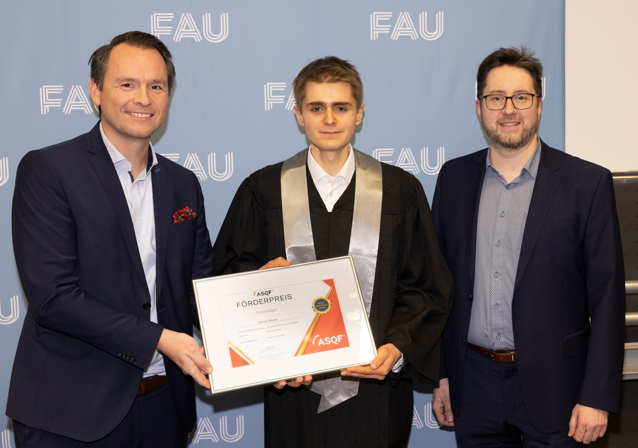 Drei Männer stehen vor einer FAU-Kulisse. Der Mann in der Mitte, der eine Abschlussrobe trägt, hält eine gerahmte Urkunde. Die beiden Männer neben ihm sind in Geschäftskleidung gekleidet und lächeln in die Kamera.