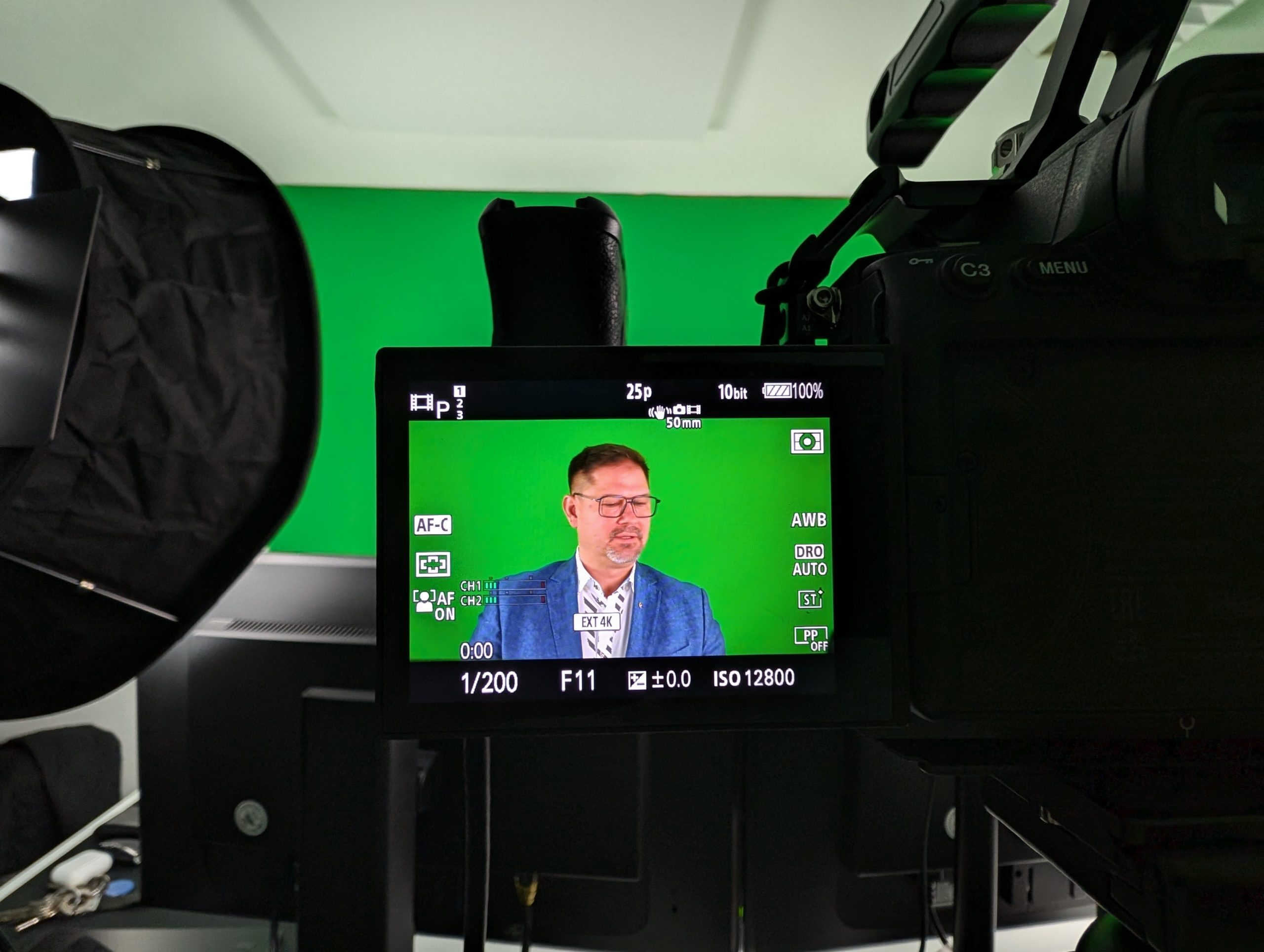 Auf einem Kamerabildschirm ist ein Mann in einem blauen Anzug zu sehen, der vor einem Greenscreen in einem Studio spricht. Um ihn herum sind professionelle Beleuchtungs- und Aufnahmegeräte zu sehen.