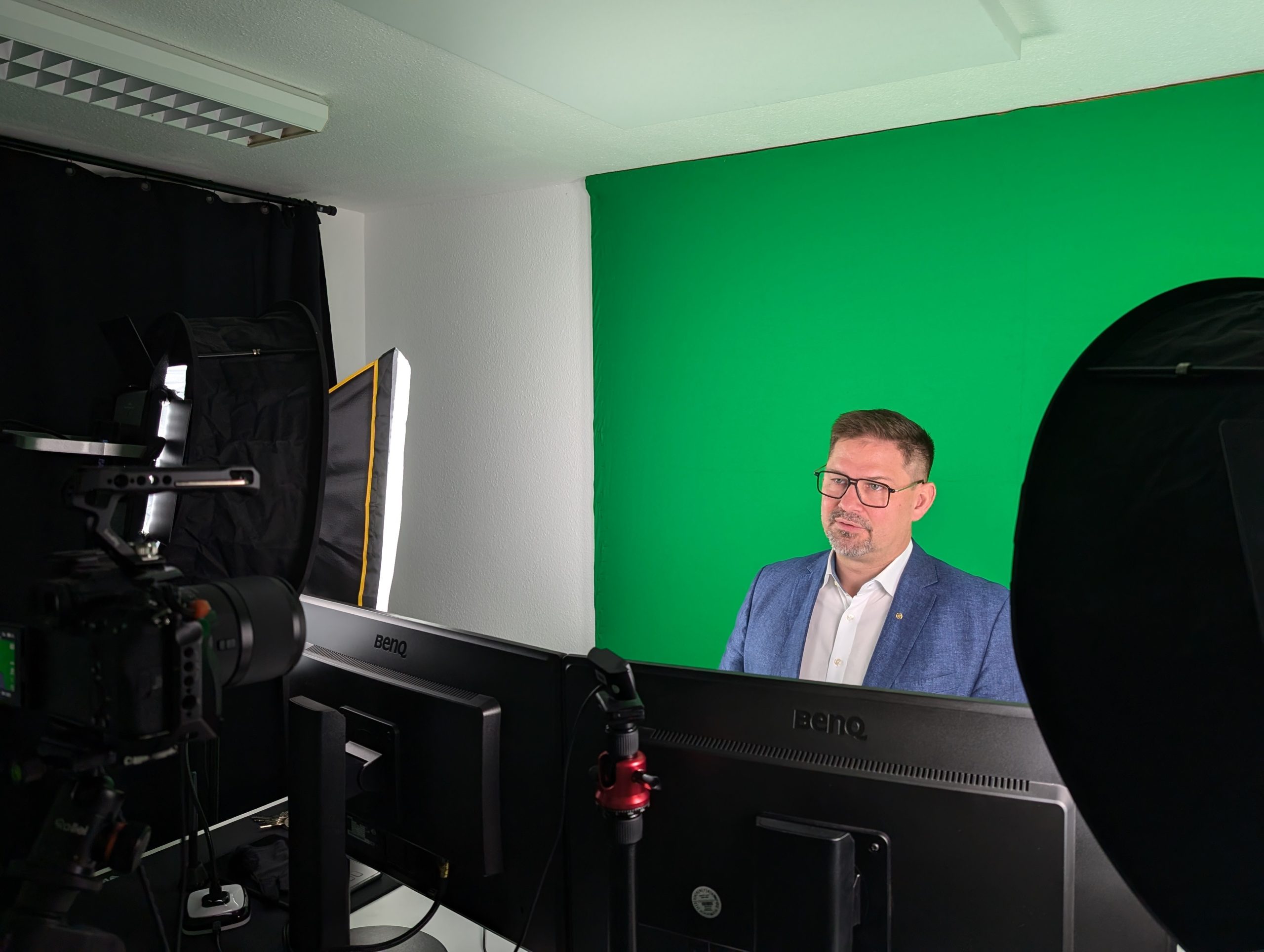 Ein Mann in einem blauen Blazer sitzt vor einem grünen Bildschirm, umgeben von Studiolampen, Monitoren und einer Kameraausrüstung und bereitet sich auf eine Videoaufnahme oder einen Livestream vor.