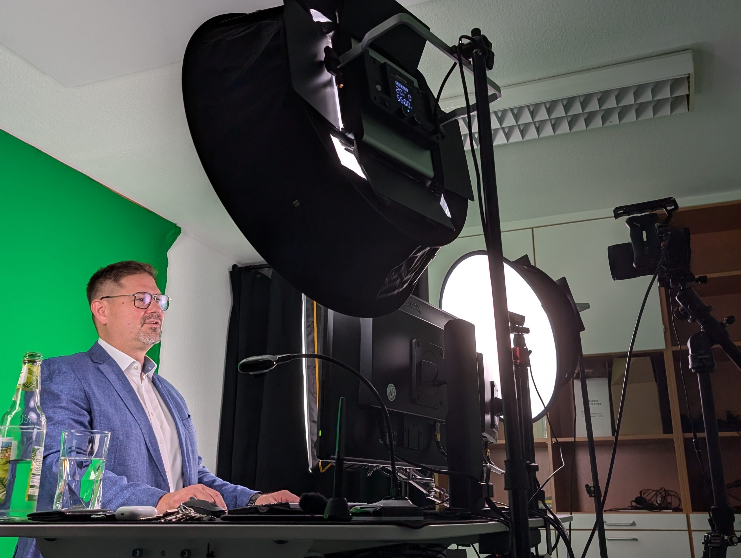 Ein Mann in einem blauen Blazer sitzt an einem Schreibtisch mit einem Computer vor einem Greenscreen und großen Studiolampen und bereitet sich auf eine Videoaufnahme oder einen Livestream vor. Auf dem Schreibtisch sind auch eine Kamera und eine Wasserflasche zu sehen.