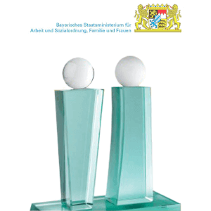 Zwei gläserne Auszeichnungen mit kugelförmigen weißen Spitzen stehen auf einem Glassockel. Darüber befinden sich das bayerische Wappen und der Text Bayerisches Staatsministerium für Arbeit und Sozialordnung, Familie und Frauen.