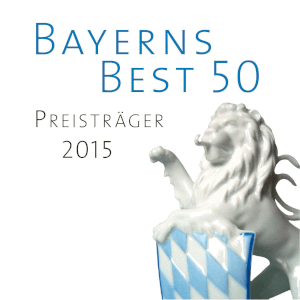 Ein weißer Porzellanlöwe mit einem blau-weiß karierten Schild vor sich. Der Text darüber lautet Bayerns Best 50 Preisträger 2015 auf weißem Grund.
