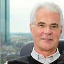 Ein älterer Mann mit weißem Haar und Brille sitzt in einem Bürostuhl und lächelt, während durch das Fenster hinter ihm eine Stadtlandschaft und ein hohes Gebäude zu sehen sind.