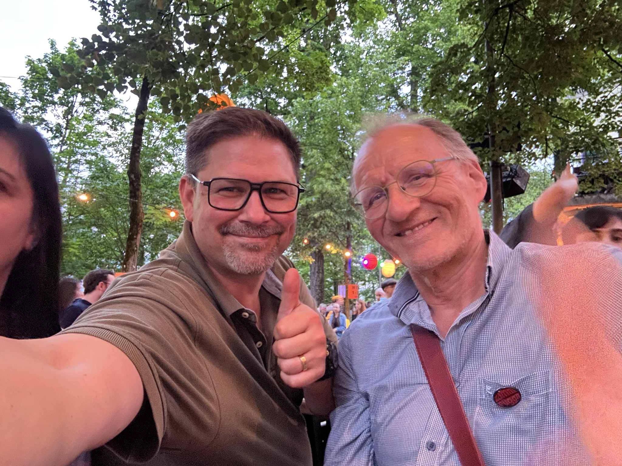 Zwei lächelnde Männer posieren bei einer lebhaften Veranstaltung im Freien für ein Selfie, wobei einer der beiden den Daumen nach oben streckt. Im Hintergrund sind bunte Lichter und Bäume zu sehen.