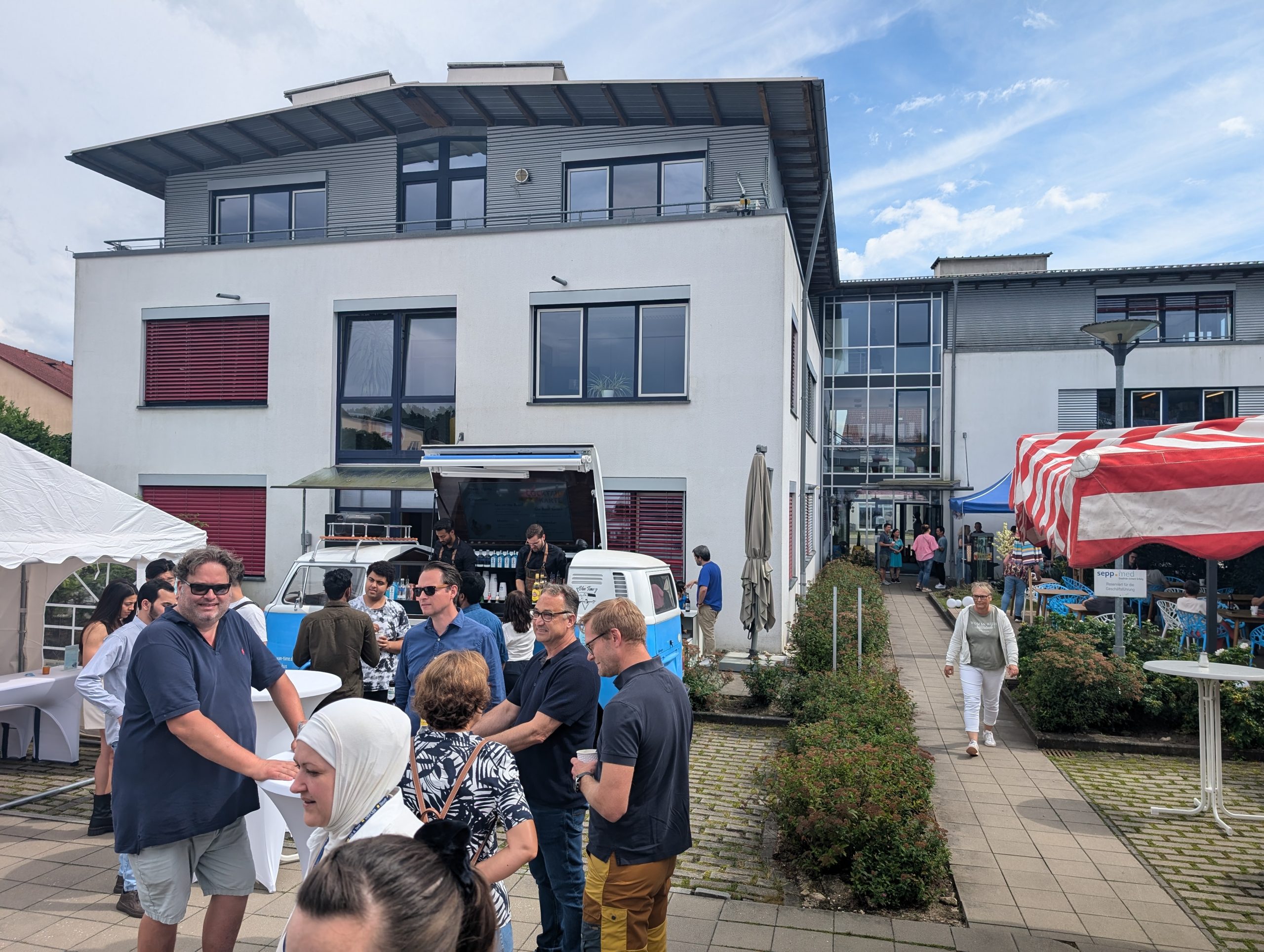 Eine Gruppe von Menschen versammelt sich und unterhält sich im Freien vor einem modernen weißen Gebäude mit Marktzelten und einem Imbisswagen. Einige Leute stehen in einer Reihe, und der Himmel ist teilweise bewölkt.