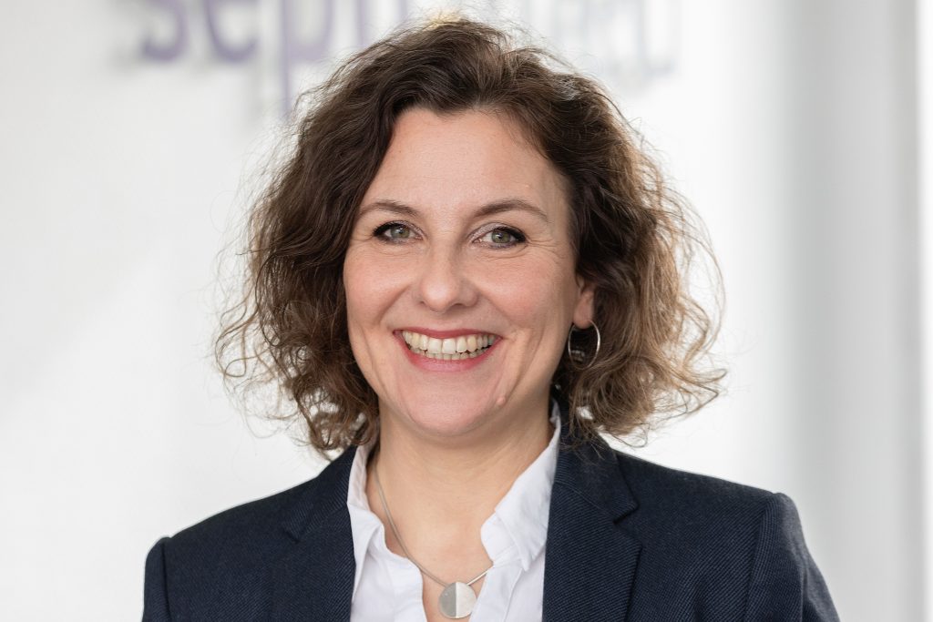 Eine Frau mit lockigem braunem Haar, die einen dunklen Blazer über einer weißen Bluse trägt, lächelt in die Kamera. Sie steht in einem hellen Innenraum mit einem unscharfen Hintergrund.