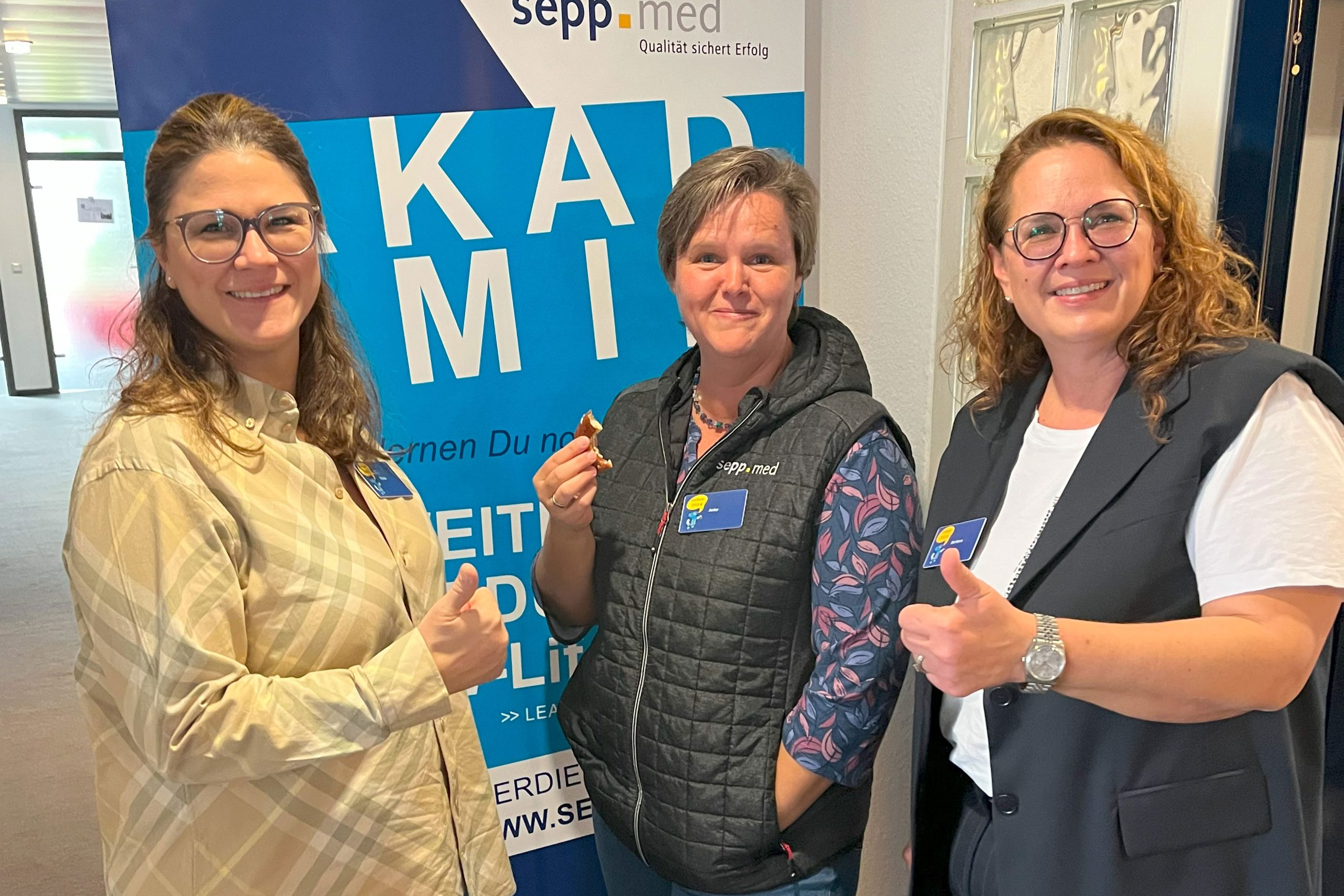 Drei Frauen, die lächeln und den Daumen hoch zeigen, stehen vor einem blau-weißen sepp.med-Banner. Sie tragen Namensschilder und legere Geschäftskleidung in einem gut beleuchteten Innenraum.