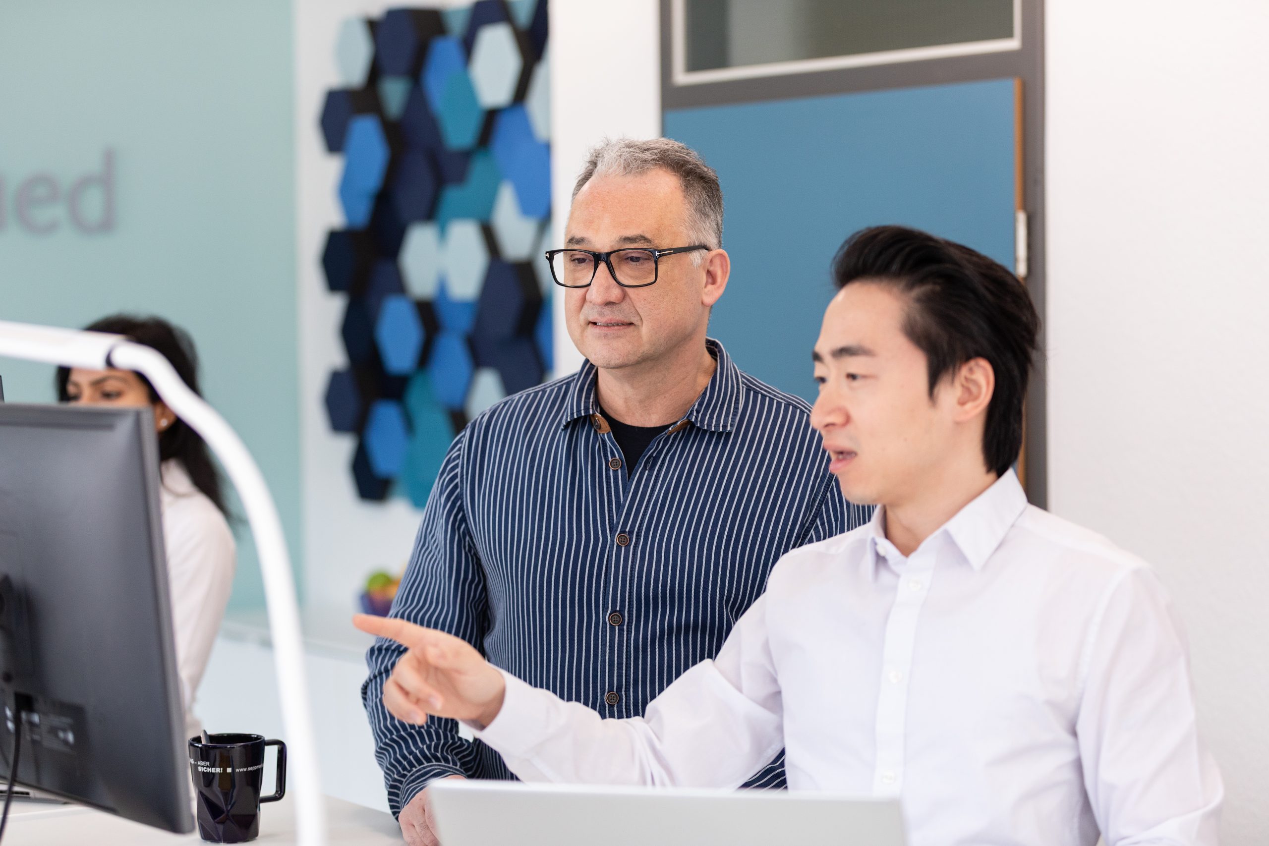 Zwei Männer arbeiten in einem Büro gemeinsam an einem Computer. Der eine, sitzend, zeigt auf den Bildschirm, während der andere, daneben stehend, aufmerksam zusieht. Im Hintergrund sind geometrische Wandkunst und eine Tür zu sehen.