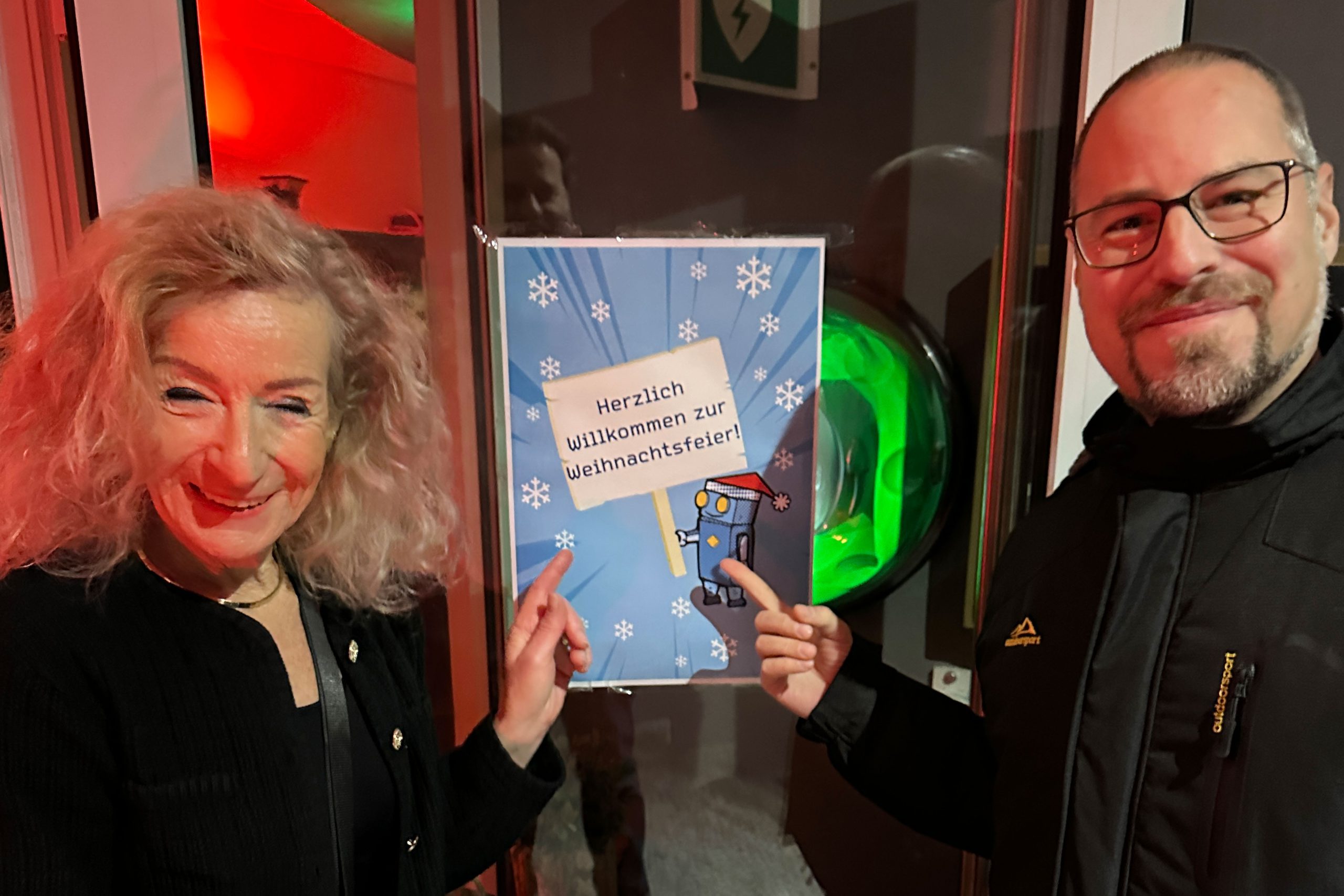 Zwei lächelnde Erwachsene stehen neben einem festlichen Schild mit der Aufschrift Herzlich Willkommen zur Weihnachtsfeier! mit Schneeflocken und einer Zeichentrickfigur in einem warm beleuchteten Raum.